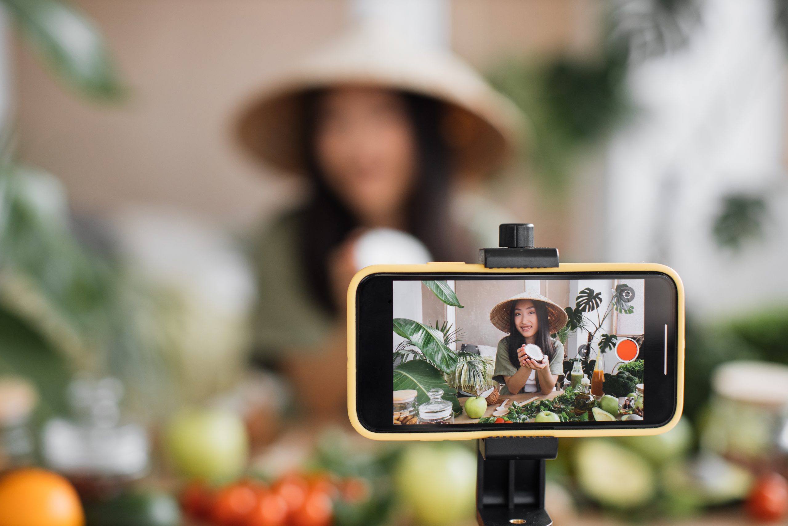 Focus on smartphone. Beauty asian blogger in traditional conical hat using coconut to make smoothies for breakfast and recording video for online channel live.
